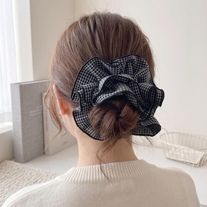 May include: A black and white checked scrunchie in a bun hairstyle. The scrunchie is made of a ruffled fabric, a fashionable hair accessory. The hair is brown and the person is wearing a white sweater.
