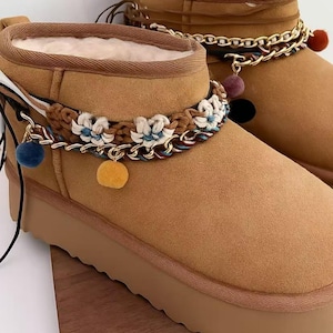 Puede incluir: Botines color canela con detalles florales y pompones decorativos. Las botas tienen una suela de plataforma y un forro de piel sintética suave. Una banda trenzada con detalles florales y pompones colgantes adorna el tobillo.