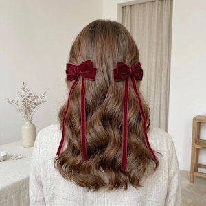 Puede incluir: Dos lazos de terciopelo rojo oscuro para el cabello unidos a cintas de terciopelo largas y delgadas. Cada lazo está sujeto a cada lado de la cabeza, con las cintas colgando verticalmente sobre el cabello castaño ondulado. Diseño clásico.