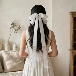 Puede incluir: Una mujer con cabello oscuro largo lleva un vestido blanco y un gran lazo blanco en el pelo. El lazo tiene cintas largas y fluidas. El fondo es de color neutro con un sofá y objetos decorativos.