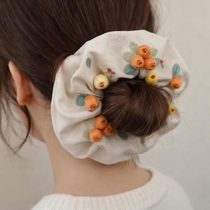 Peut inclure: Un chouchou beige avec des broderies de grappes de baies orange et jaunes et de feuilles vertes. Le chouchou est enroulé autour d'un chignon, avec le dos de la tête et du cou d'une personne visible.