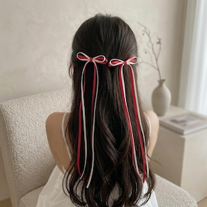 May include: A person's dark brown hair styled with two small bows. Each bow is red and white, with long matching ribbons cascading down the back. The person is wearing a white top, and the background is neutral.