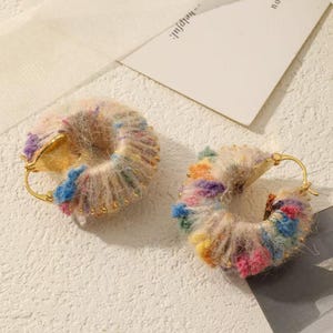 May include: A pair of gold-tone hoop earrings. Each earring is decorated with a fluffy, multi-coloured material, featuring pink, blue, yellow, and purple hues. Tiny gold-coloured beads are set along the outer edge of the hoops.
