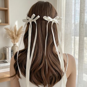 Könnte beinhalten: Nahaufnahme des Hinterkopfes einer Person mit langen, welligen, braunen Haaren. Zwei weiße Satin-Schleifen sind im Haar befestigt, jede mit langen, fließenden Bändern. Die Bänder fallen über den Rücken und erzeugen einen zarten Look.