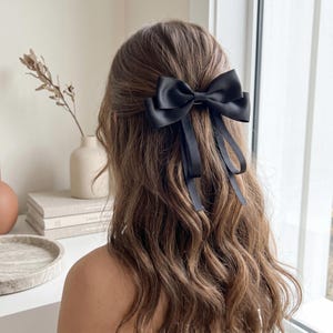 Könnte beinhalten: Eine schwarze Satin-Haarschleife ist in welligem braunem Haar befestigt. Die Schleife ist im klassischen Stil mit langen, fließenden Bändern gebunden. Das Haaraccessoire verleiht der Frisur einen Hauch von Eleganz und Raffinesse.