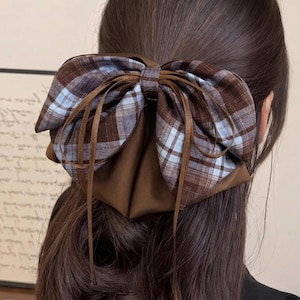 Könnte beinhalten: Eine braune Haarschleife mit einem Karomuster in Braun- und Hellblautönen. Die Schleife ist mit braunen Bändern gebunden und vor dem Hintergrund von dunkelbraunem Haar platziert.