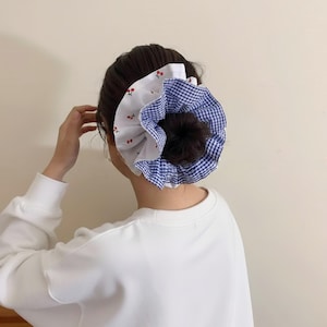 May include: A large scrunchie in a bun hairstyle. The scrunchie features a white fabric with red cherry prints and a blue and white gingham pattern. The person is wearing a white long-sleeved shirt.