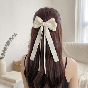 Peut inclure: Un nœud de cheveux crème avec de longs rubans flottants est attaché à l'arrière de la tête. Le nœud est centré et ajoute une touche d'élégance. Les cheveux sont longs et bruns, avec une légère ondulation.
