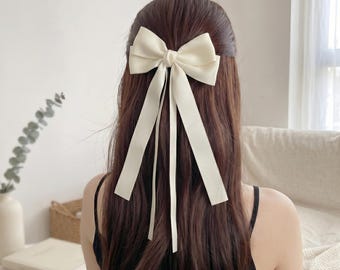 May include: A cream-colored hair bow accessory featuring a structured bow at the top with two long, flowing ribbon tails extending downward. The smooth, solid-colored fabric is secured to the hair, creating a clean and simple decorative accent.