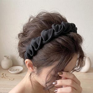 Puede incluir: Diadema de tela negra con diseño plisado y finas rayas blancas. La diadema se lleva en la cabeza de una persona, con cabello castaño oscuro visible. La diadema es un elegante accesorio para el cabello.