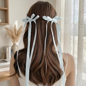 Puede incluir: Dos lazos de pelo de cinta azul claro están sujetos al cabello castaño. Los lazos están atados en forma de lazo y tienen cintas largas y fluidas. El cabello está peinado con ondas sueltas. El fondo es de color neutro.