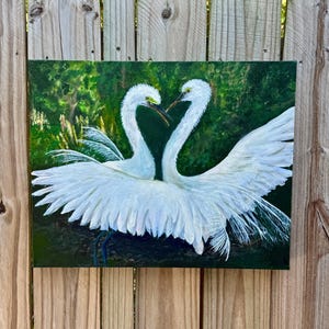 以下が含まれることがあります： 白いサギ2羽が首を絡ませてハート型を作った絵画。鳥は緑豊かな背景を背景に翼を広げています。このアートワークは、静けさと自然の美しさを呼び起こします。