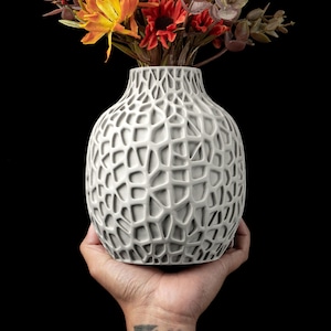 Peut inclure: Un vase en céramique grise avec un design unique en cellules ouvertes, rempli de fausses fleurs colorées. Le vase est tenu dans une main sur un fond noir. Les fleurs comprennent des teintes orange, rouge et verte, ajoutant un contraste vibrant au vase.