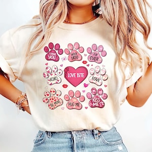 May include: A cream-coloured t-shirt with pink paw prints and Valentine's Day messages. The phrases include "I chews you," "Woof you," "Fur ever," "Love bite," "XOXO," "Be mine," "Hug me," and "Kiss me."