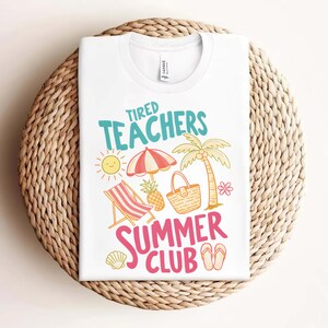 May include: White t-shirt with the text "Tired Teachers Summer Club" in teal and pink. The graphic includes a sun, beach umbrella, palm tree, beach chair, pineapple, and flip-flops. The shirt is folded on a woven surface.