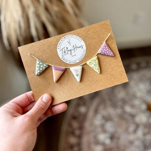 Puede incluir: Una pancarta de arcilla hecha a mano con cinco banderines triangulares en varios colores y patrones, ensartados en hilo. Los banderines presentan diseños como corazones, lunares y motivos florales. La pancarta se muestra en una tarjeta marrón con el texto "T. Klay Haus".