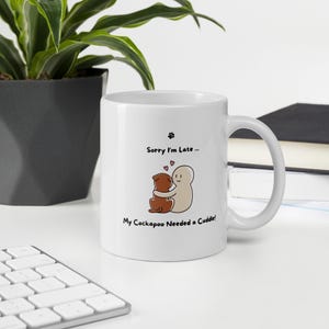 Pode incluir: Caneca de cerâmica branca com uma alça. A caneca apresenta uma ilustração de desenho animado de um cão abraçando uma pessoa, com o texto "Sorry I'm Late... My Cockapoo Needed a Cuddle!" acima e abaixo da ilustração. Uma pegada está no topo.