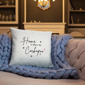 以下が含まれることがあります： 「Home is where the Cockapoo is」という黒い文字が書かれた白い正方形のクッション。 「Cockapoo」という単語は大きなフォントサイズです。 小さな足跡とピンクのハートがテキストを強調しています。 クッションは青いニットの毛布の上にあります。