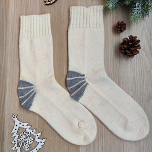 May include: A pair of cream-colored knit socks with gray heels and toes. The socks have ribbed cuffs and are displayed on a wooden surface with decorative pine cones and a wooden Christmas tree ornament.