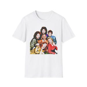 May include: White t-shirt featuring a group portrait of six people. The image on the shirt shows a group of people with various hairstyles and clothing. The shirt is made of a soft, comfortable material.