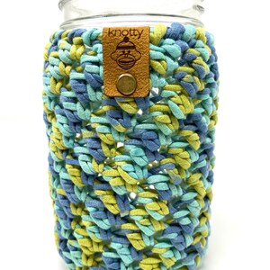 May include: A clear glass jar with a blue, green, and yellow crocheted cozy. The cozy has a brown leather tag with the word "knotty" and a monkey logo. The cozy is designed to fit around the jar.