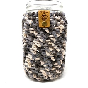 May include: A clear glass jar with a crocheted cozy in shades of gray and cream. The cozy has a textured, woven appearance. A small, brown leather tag with the word "knotty" and a monkey logo is attached to the jar.