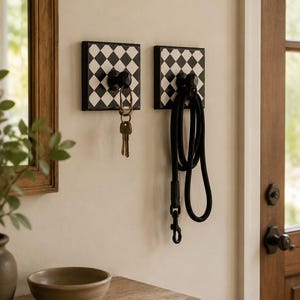 May include: Two square wall-mounted hooks featuring a black and white checkered pattern. Each hook has a central black knob. One hook holds a set of metal keys, while the other hook holds a black rope leash with a metal clip attachment.