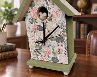 May include: A house-shaped clock with a sage green wooden frame and a white front panel featuring a floral pattern of pink, peach, and green leaves. A small white bird figure sits in the circular opening above the black clock hands.