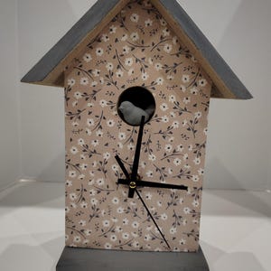 May include: A decorative birdhouse clock with a beige floral pattern and a gray roof and base. The clock features black hands and a small bird figure in the circular opening. The clock is a unique home decor item.