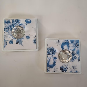 May include: Two square cabinet knobs with a white and blue floral design. Each knob has a clear, faceted crystal handle. The knobs are set against a white background.