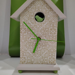May include: A decorative birdhouse clock with a cream-colored wooden frame and a patterned face. The clock features bright green clock hands and a small green bird inside. The base and roof are painted cream, and the clock sits on small green feet.