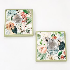 May include: Two square wooden wall hooks with a light green frame and a floral pattern featuring pink, orange, and blue flowers on a white background. Each hook is centered with a clear, faceted, diamond-shaped glass knob for hanging items.