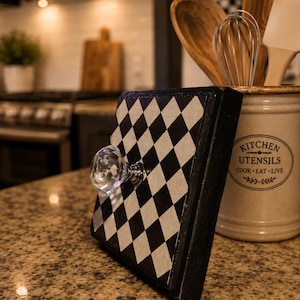 May include: A square wooden sign featuring a black and white diamond harlequin pattern on the front face. A clear, faceted crystal-style knob is attached to the center of the sign. The edges of the wooden block are painted in a solid black finish.