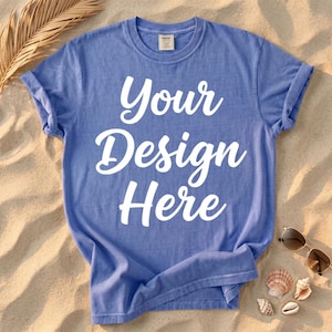 以下が含まれることがあります： 袖をまくった青いTシャツに、白い文字で「Your Design Here」と書かれています。 砂浜の上に、サングラス、貝殻、乾燥したヤシの葉と一緒に置かれています。
