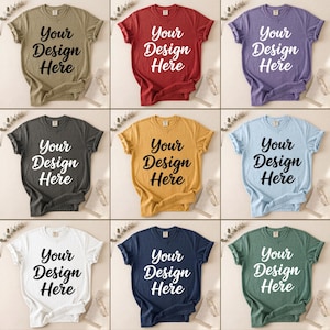 以下が含まれることがあります： 「Your Design Here」のテキストが太字の筆記体で書かれた、9色の異なる色のTシャツ。色はオリーブグリーン、赤、紫、チャコール、マスタードイエロー、ライトブルー、白、ネイビーブルー、フォレストグリーンです。