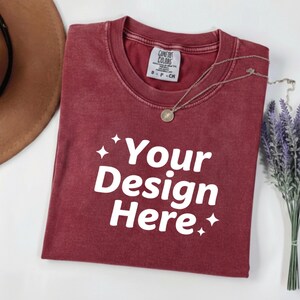 May include: A maroon t-shirt with the text "Your Design Here" in white. The shirt has a crew neck and a small tag on the collar. A silver necklace with a pendant and a sprig of lavender are also visible.
