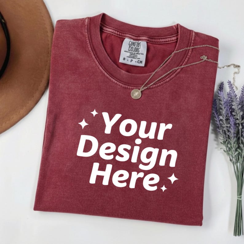 May include: A maroon t-shirt with the text "Your Design Here" in white. The shirt has a crew neck and a small tag on the collar. A silver necklace with a pendant and a sprig of lavender are also visible.