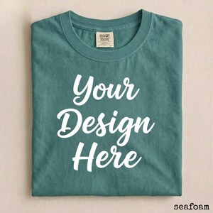 May include: Seafoam green t-shirt with the text "Your Design Here" in white script. The shirt is folded neatly, showcasing the design. A small tag is visible near the collar. The word "seafoam" is in the bottom right corner.