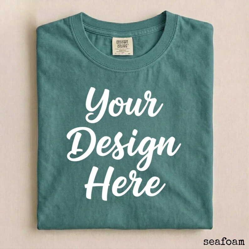 May include: Seafoam green t-shirt with the text "Your Design Here" in white script. The shirt is folded neatly, showcasing the design. A small tag is visible near the collar. The word "seafoam" is in the bottom right corner.