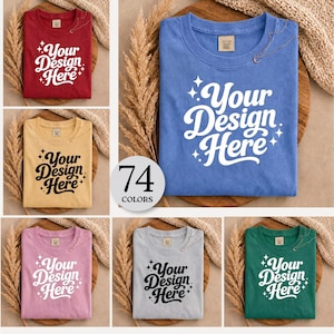 Peut inclure: Plusieurs t-shirts pliés de différentes couleurs : rouge, jaune, bleu, rose, gris et vert. Chaque t-shirt affiche le texte "Your Design Here" dans une police stylisée. Un graphique circulaire indique "74 COLORS".