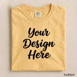 May include: A folded, butter-colored t-shirt with the words "Your Design Here" in a black, cursive font. The shirt has a crew neck and a small tag at the neckline. The word "butter" is in the bottom right corner.