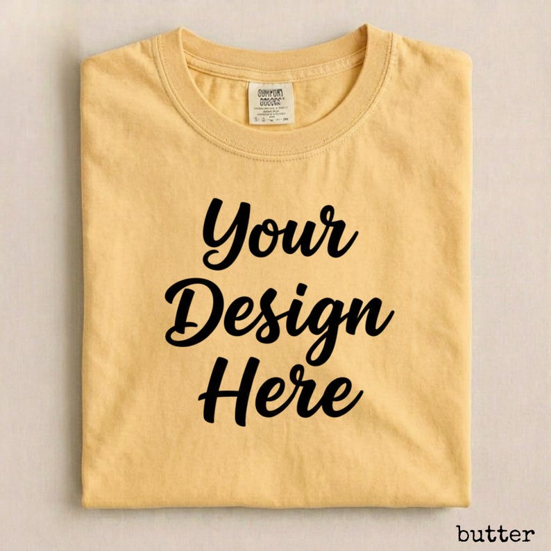 May include: A folded, butter-colored t-shirt with the words "Your Design Here" in a black, cursive font. The shirt has a crew neck and a small tag at the neckline. The word "butter" is in the bottom right corner.