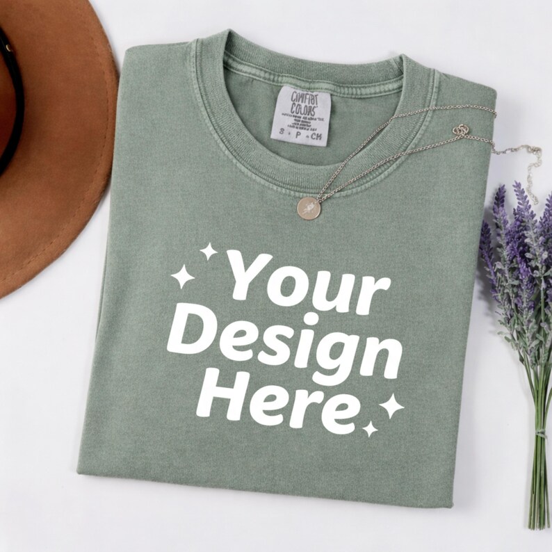 May include: Sage green t-shirt with the text "Your Design Here" in white. A silver necklace with a round pendant rests on the shirt. A brown hat and a bundle of lavender are also in the image.