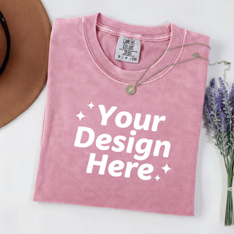 May include: A dusty rose t-shirt with the text "Your Design Here" in white. A silver necklace with a circular pendant rests on the shirt. A brown hat and a bundle of lavender are in the background. The shirt has a tag that says "Comfort Colors".