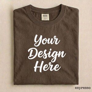 May include: Espresso-colored t-shirt with the text "Your Design Here" in white script. The shirt is folded neatly, showcasing the design. A tag is visible near the collar. The shirt is a blank canvas for custom designs.