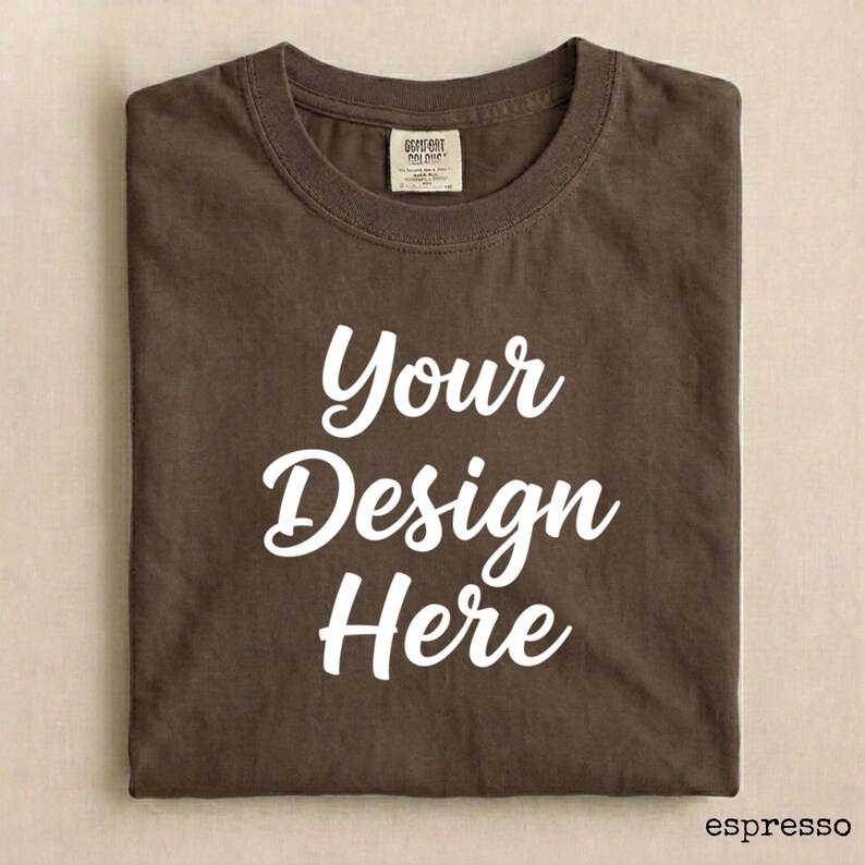 May include: Espresso-colored t-shirt with the text "Your Design Here" in white script. The shirt is folded neatly, showcasing the design. A tag is visible near the collar. The shirt is a blank canvas for custom designs.