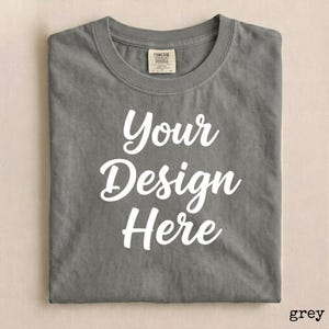 May include: A grey t-shirt with the words "Your Design Here" in white script. The shirt has a crew neck and a tag at the neckline. The shirt is folded neatly.