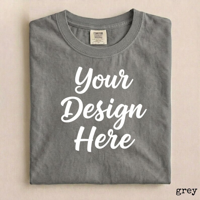 May include: A grey t-shirt with the words "Your Design Here" in white script. The shirt has a crew neck and a tag at the neckline. The shirt is folded neatly.