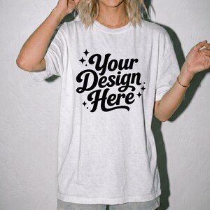 Pode incluir: Uma t-shirt branca de manga curta com o texto preto Your Design Here numa fonte cursiva arrojada. O design &eacute; acentuado com pequenos &iacute;cones de estrelas de quatro pontas que rodeiam as letras, criando uma apresenta&ccedil;&atilde;o gr&aacute;fica limpa.
