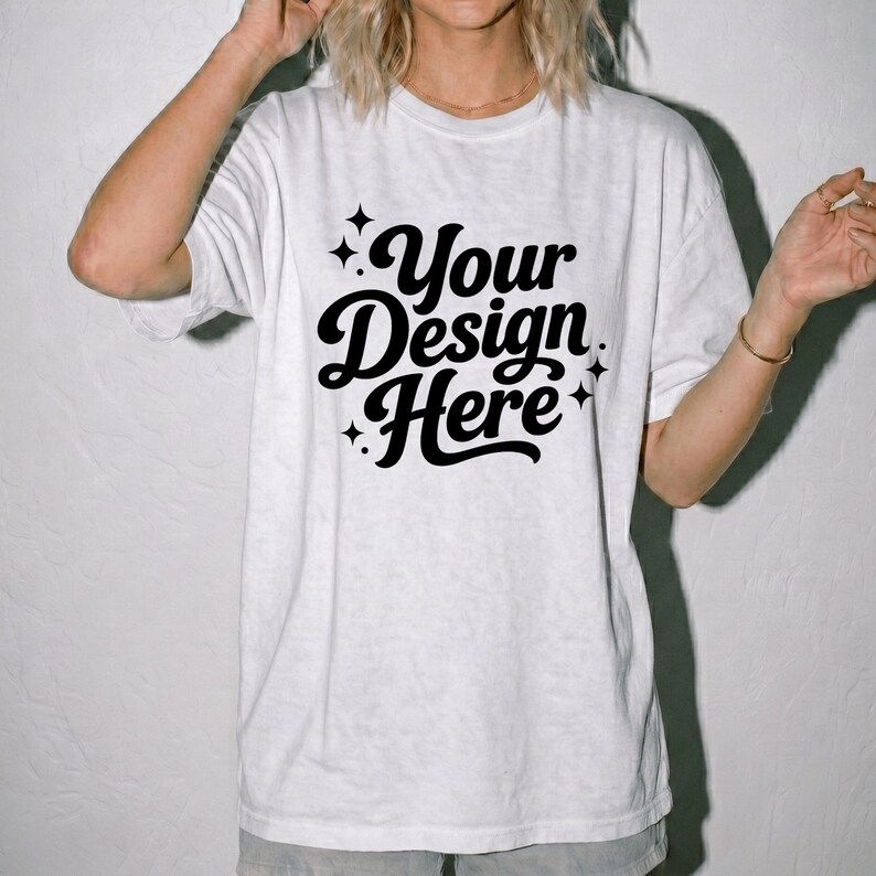 Pode incluir: Uma t-shirt branca de manga curta com o texto preto Your Design Here numa fonte cursiva arrojada. O design &eacute; acentuado com pequenos &iacute;cones de estrelas de quatro pontas que rodeiam as letras, criando uma apresenta&ccedil;&atilde;o gr&aacute;fica limpa.